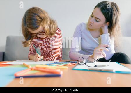 Young female teacher giving private lesson to child, little girl sitting at her desk writing in notebook. Stock Photo