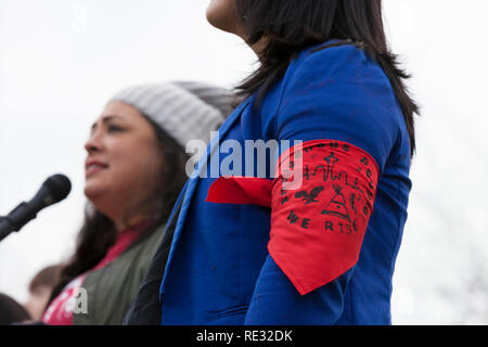 Washington, USA. 19th Jan 2019.  Monserrat Padilla, coordinator at the Washington Immigrant Solidarity Network and transgender advocate, wears an armband in support of all indigenous peoples at the pre-march rally for the Womxn's March Seattle 2019.  Speaking is Colleen Echohawk, member of the Kithehaki Band of the Pawnee Nation. Saturday’s March and Rally, organized by Seattle Womxn Marching Forward in collaboration with MLK Day organizers, will be followed on Sunday by a day of action and Monday MLK Day events honoring Martin Luther King Jr. Sunday’s Womxn Act on Seattle is a citywide day of Stock Photo