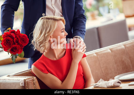 Young couple on date in restaurant man standing with bouquet behind sitting woman holding his hand cheerful Stock Photo