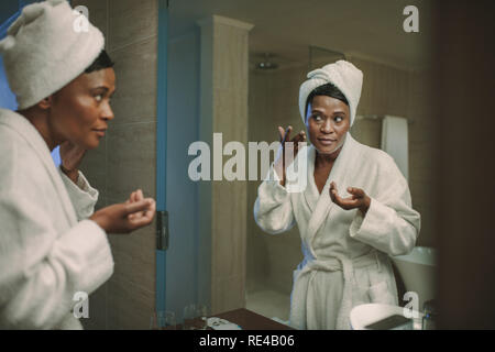 African woman applying cream in bathroom mirror after her bath. Woman taking care of her skin and applying face cream in bathroom. Stock Photo