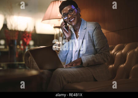 Happy mature businesswoman sitting in hotel lobby and talking on mobile phone with laptop. Businesswoman on trip working from hotel lobby. Stock Photo