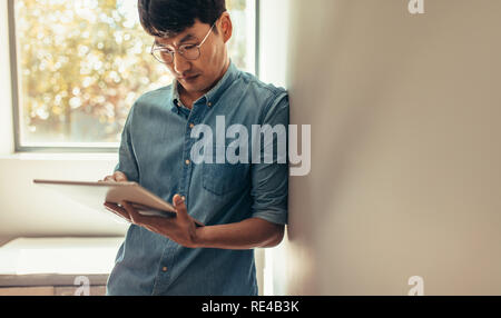 Asian man leaning to a wall and using digital tablet. Man in casuals standing at home using tablet computer to update his social media status. Stock Photo