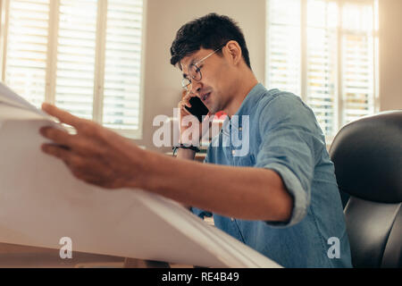 Male interior designer looking at drawing and talking over smart phone. Architect using phone at his workplace. Stock Photo