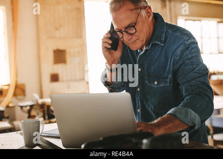 Mature man looking busy using laptop and talking on mobile phone in his workshop. Senior male carpenter working on laptop and phone call in his carpen Stock Photo