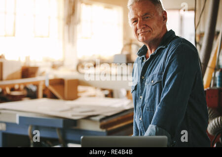 Portrait of confident senior male carpenter standing in his workshop. Mature male carpenter looking at camera. Stock Photo