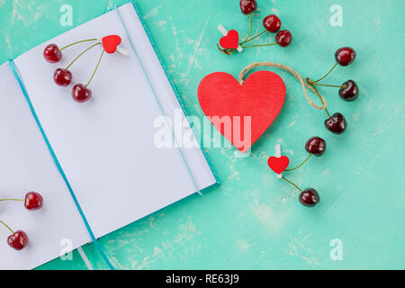 Composition with diary and sweet cherries and pen, open white paper note book and decor with red cherry, wooden heart on green wooden table.love diary and red hearts Stock Photo