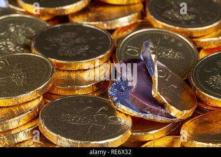 Chocolate money pile of gold coins. Edible sweets covered in foil as pretend money. One open coin half eaten Stock Photo