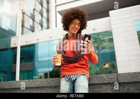 A handsome curly-haired guy of mixed race is dressed in a trendy youth-style scarf and sweater writes a message in a teleone against the background of Stock Photo