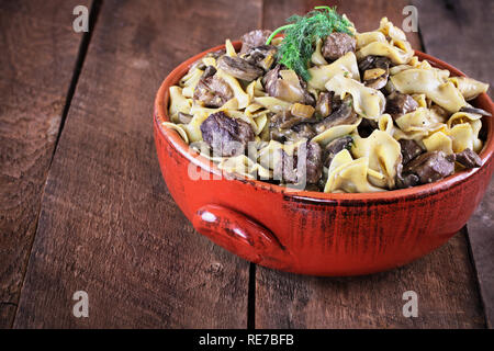 Venison or Beef Mushroom Stroganoff with in a red ceramic serving bowl garnished with fresh dill over a rustic wood table background. Stock Photo
