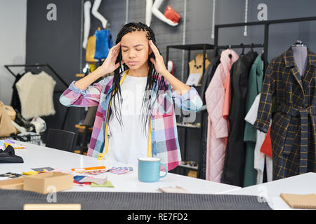 Young female tailor holding head in her hands, she has a headache, while she working over new fashion collection in workshop Stock Photo