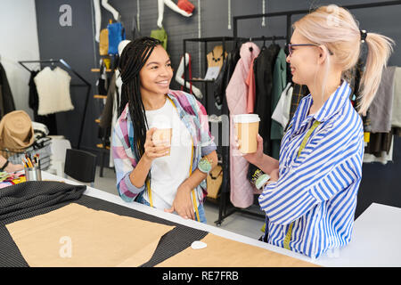 Two young female tailors smiling and talking to each other while standing and drinking coffee in workshop Stock Photo