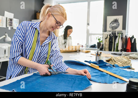 Young female tailor in eyeglasses cutting pattern on fabric with scissors with her colleague working in the background Stock Photo