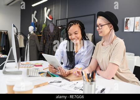 Fashion designers sitting at the table among sketches and palettes and using digital tablet together in workshop Stock Photo