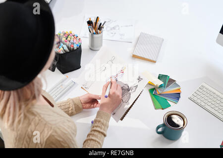 Rear view of young designer in hat sitting at the table and drawing fashion sketches on paper Stock Photo