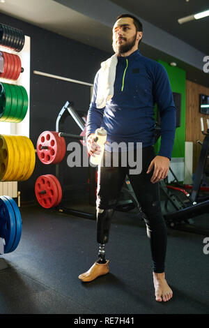 Disabled bearded young man in sportswear standing with bottle of water in gym Stock Photo