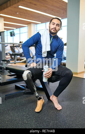 Portrait of young disabled man with amputee leg sitting with bottle of water and resting after sport training in gym Stock Photo