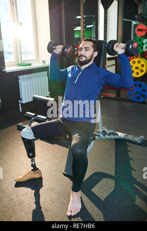 Content handsome young bearded man with amputated leg sitting on sport bench and lifting small weights while training in gym Stock Photo