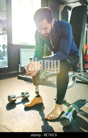 Sad amputee young bearded man sitting on sort bench and leaning on knees in health club, small dumbbells on floor Stock Photo