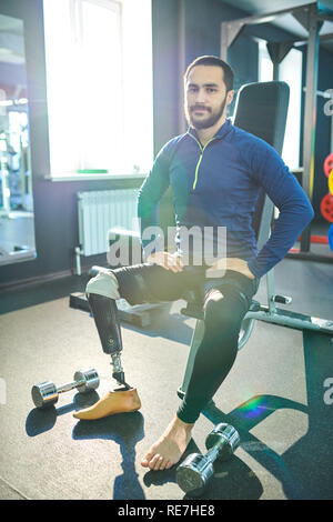 Content confident handsome young bearded man with prosthetic leg sitting on sport bench and looking at camera in modern health club Stock Photo