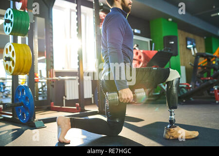 Serious athletic young man with prosthetic leg standing on one knee and holding dumbbells while performing exercise with weight in gym Stock Photo