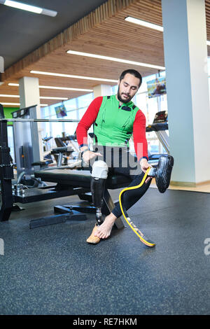 Serious handsome young bearded man with amputated leg sitting on sport bench and changing prosthesis while preparing for fitness training Stock Photo