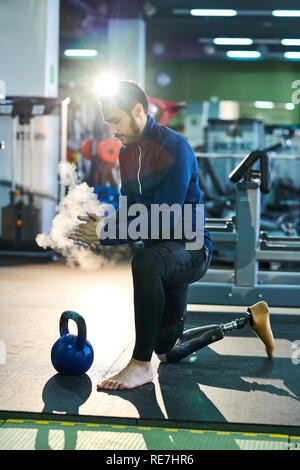Serious handsome young bearded man with prosthetic leg standing on one knee and clapping hands with talc powder while ready to exercise with kettlebel Stock Photo