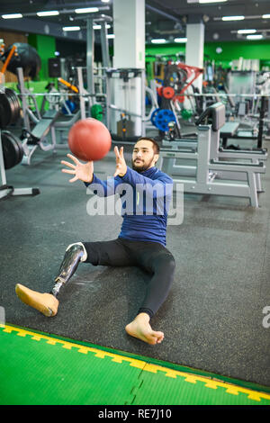 Content handsome young bearded man with prosthetic leg sitting on floor in modern gym with fitness equipment and catching ball Stock Photo