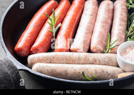 Close-up assorted fresh raw sausages with thyme, rosemary and garlic in a cast iron pan on a dark concrete table. Stock Photo