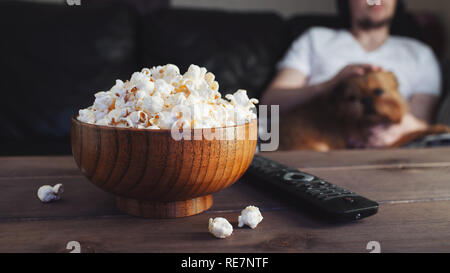 Wooden bowl with salted popcorn and TV remote on wooden table. In the background, a man with a red dog watching TV on the couch. Stock Photo