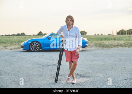 The beautiful young brutal man stands at a blue sports car at sunset, he is dressed in a white shirt with a short sleeve and red shorts Stock Photo