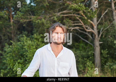 Portrait of the young handsome brutal man with long hair, the blue eyes, an easy bristle, he is dressed in a white shirt with a short sleeve Stock Photo