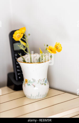 Closeup of old-fashioned ceramic porcelain flower vase with yellow plants and landline phone on wooden table in room corner Stock Photo