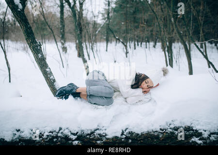 Young sad woman is lying and resting on the snow covered trunk of tree during the walk in the winter forest. Stock Photo