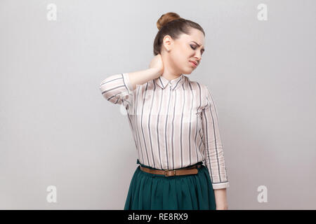 Neck pain. Portrait of unhappy beautiful young woman in striped shirt and green skirt and collected ban hairstyle, standing and holding her painful ne Stock Photo