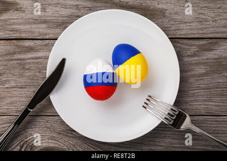 Two Easter eggs, painted in color of the flags of Russia and Ukraine in a white plate on wooden table, top view Stock Photo