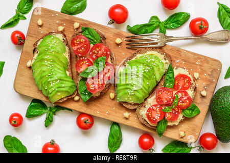 sandwiches with avocado, tomatoes, basil, hummus and sesame seeds on rustic cutting board over white marble table. Healthy breakfast. top view Stock Photo