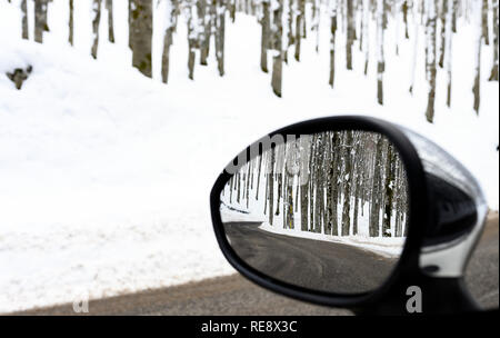 View of a road that passes in the middle of a forest of snow-covered pine trees seen through the rearview mirror of a car. Winter season in Italy. Stock Photo
