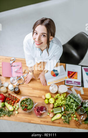 Portrait of a young woman nutritionist in medical uniform standing near the table full of various healthy products indoors Stock Photo