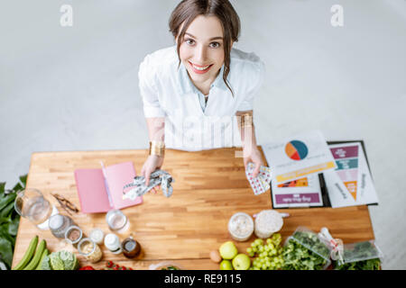 Portrait of a young woman nutritionist in medical uniform standing near the table full of various healthy products indoors Stock Photo