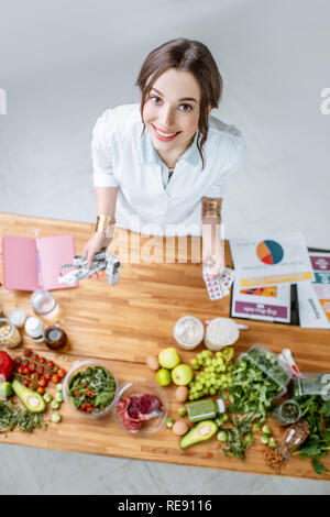 Portrait of a young woman nutritionist in medical uniform standing near the table full of various healthy products indoors Stock Photo