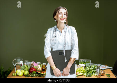Portrait of a young woman nutritionist in medical gown standing with weights and tape measure in the office with healthy food on the background Stock Photo