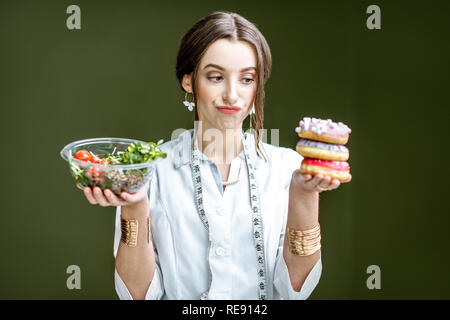 Young woman nutritionist looking on the donuts with sad emotions choosing between salad and unhealthy dessert on the green background Stock Photo