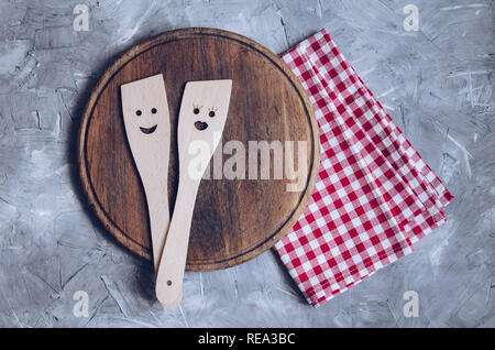 Kitchen wooden utensils with smile on red checkered napkin and round wooden board. Wooden spoons with male and female faces. Family cooking concept. R Stock Photo