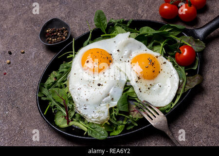 Two fried eggs, sunny side up, on pan with herbs, arugula, spinach, cherry tomatoes and pepper, with fork an brown background. Concept of healthy life Stock Photo