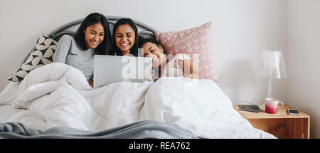 Three girls lying in bed covered in a blanket watching movie on a laptop. Girls having fun using a laptop lying on bed during a sleepover. Stock Photo