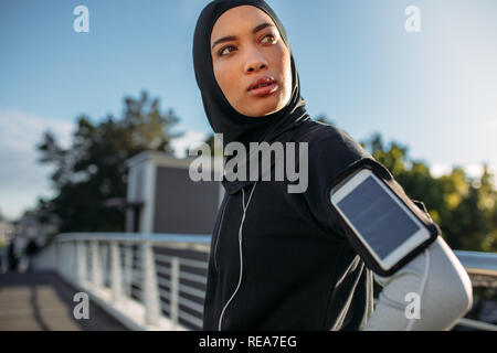 Young sportswoman in hijab standing outdoors with mobile phone on her armband looking away. Healthy female taking a break after exercising outdoors in Stock Photo