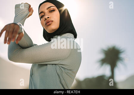 Young sportswoman in hijab stretching her arms and looking at camera. Muslim female exercising outdoors in morning. Stock Photo