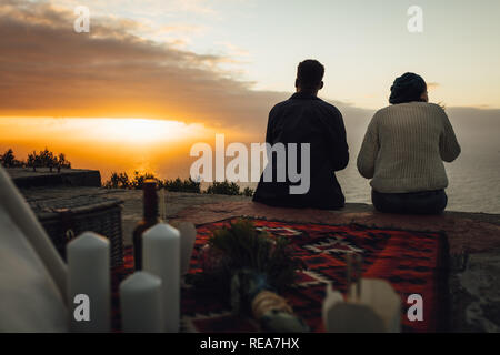 Rear view of couple sitting outdoors and looking at the sunset. Couple having picnic. Stock Photo