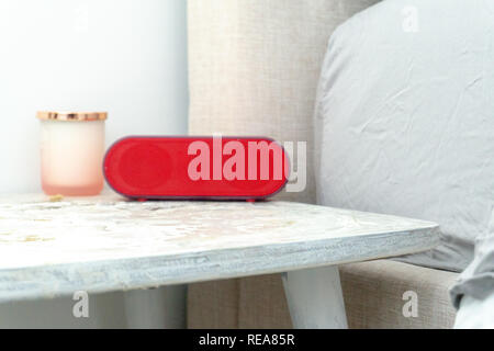 Bright red radio alarm clock and candle jar on a bedside table nightstand, with bed and linens showing in soft colors. Stock Photo