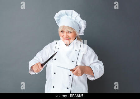 Senior woman chef studio standing isolated on gray with knives frightening playful Stock Photo